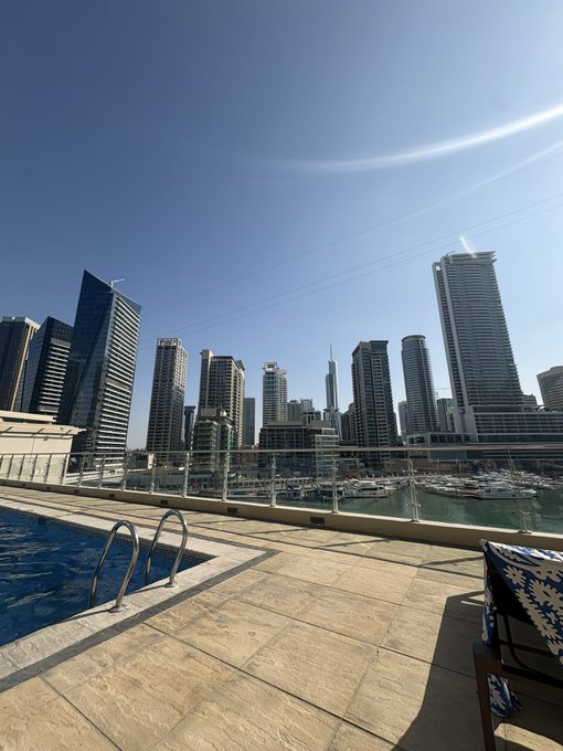 A rooftop pool overlooking a stunning marina filled with yachts and a backdrop of modern skyscrapers under a clear blue sky, paired with the post "MAY I GET A GM? 💙"—the tranquil yet vibrant urban scene radiates good morning energy, inviting viewers to start their day with a sense of luxury and positivity.
