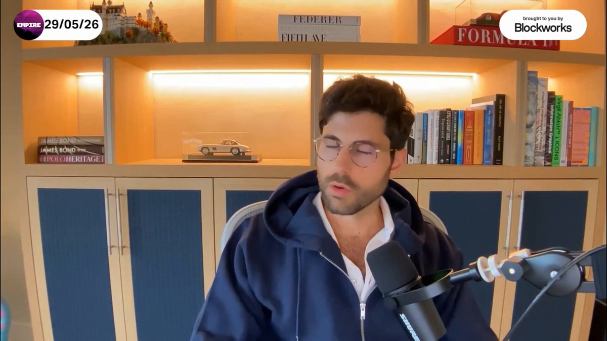 The image features a man wearing glasses sitting at a desk with a microphone, likely recording a podcast or live stream. The background includes bookshelves filled with various books, suggesting that he might be discussing topics related to finance, technology, or other subjects of interest. The man is dressed in a blue hoodie and appears to be engaged in the content being shared on his show.