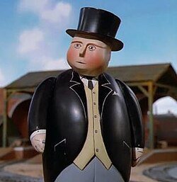 Sir Topham Hatt, the controller character from the children's show Thomas & Friends, stands in front of a railway yard wearing his signature black top hat and formal suit.