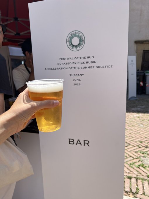 The image shows a person holding a cup filled with beer, likely at an outdoor event or festival. There are also two other people present in the scene, one standing near a sign that says "Bar" and another person who appears to be behind the sign. The background of the photo features a brick wall and some trees, giving it a casual and relaxed atmosphere.