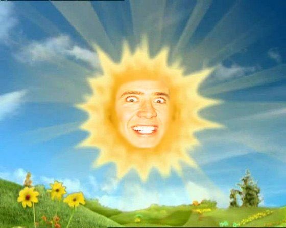 A digitally edited sun with Nicholas Cage's smiling face shines brightly over a vibrant green meadow dotted with flowers, perfectly matching the caption "It's another beautiful day to be alive" with its cheerful, uplifting atmosphere.