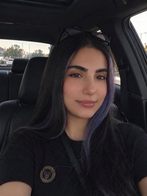 A selfie of a young woman with long dark hair featuring purple highlights, sitting in the driver's seat of a car wearing a black t-shirt with sunglasses resting on her head.