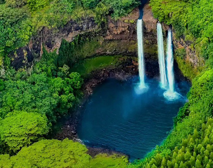 An aerial view of a lush green tropical landscape with three waterfalls cascading into a deep blue pool.
