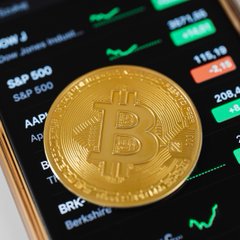 A golden Bitcoin coin rests on a smartphone displaying a stock trading app with tickers like "S&P 500" and "AAPL," visually representing the cryptocurrency's market activity discussed in the post about Bitcoin's MVRV ratio rising above its 365-day moving average.