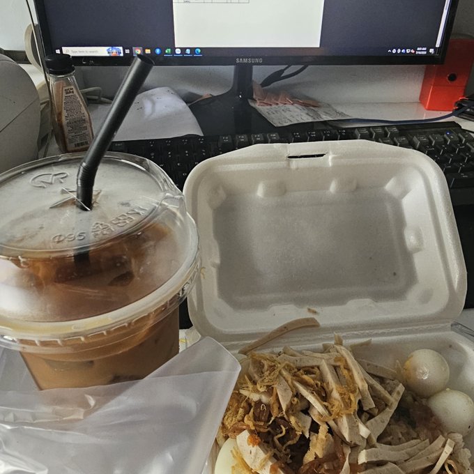 An iced coffee with a straw sits next to a takeout container of shredded chicken and boiled eggs on a cluttered desk with a Samsung monitor.