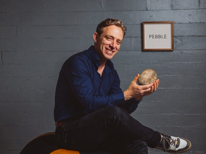 A smiling man sits against a gray wall holding a large stone, beside a framed sign reading “PEBBLE.”