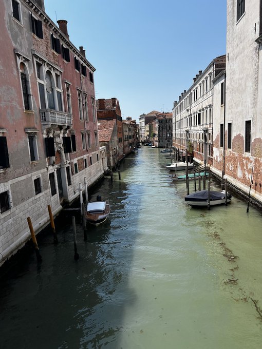 A classic Venetian canal view between aged brick and stone buildings under a clear blue sky, with a couple of small boats moored along the greenish waterway.