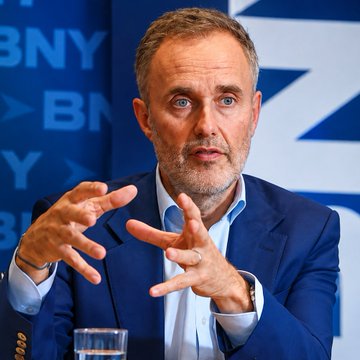 A middle-aged man with gray hair and a beard, wearing a blue suit jacket, gestures with his hands while speaking at what appears to be a BNY (Bank of New York) event.