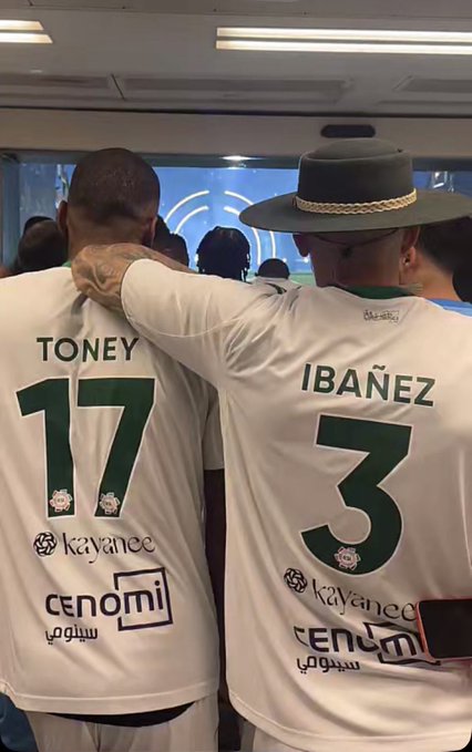 Two soccer players from behind wearing white jerseys reading 'TONEY 17' and 'IBAÑEZ 3' with 'Cenomi' sponsorship, arms around each other.