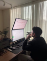 A person sits at a desk in front of a computer monitor displaying code or charts, with bright window light streaming in from behind. The image visually represents the story of a student using AI tools like Claude to develop a quantitative trading bot for Polymarket, working alone at his computer setup.
