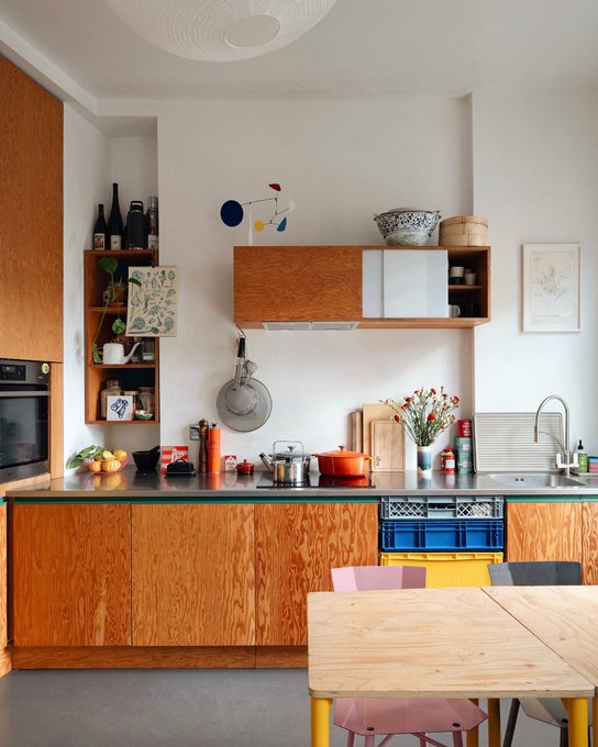A modern kitchen features warm wood cabinetry, a green countertop, a stainless steel sink, colorful chairs at a wooden table, and various cooking items including a red Le Creuset pot and fresh flowers.