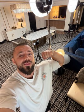 A man with a beard wearing a white t-shirt takes a selfie in a room set up with photography lighting equipment and a tripod.