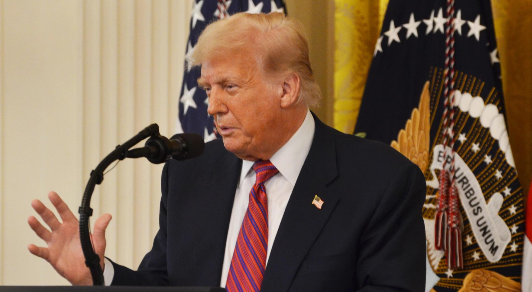 Donald Trump, wearing a dark suit with a red and blue striped tie and American flag pin, speaks at a podium with microphones in front of the Presidential Seal backdrop.