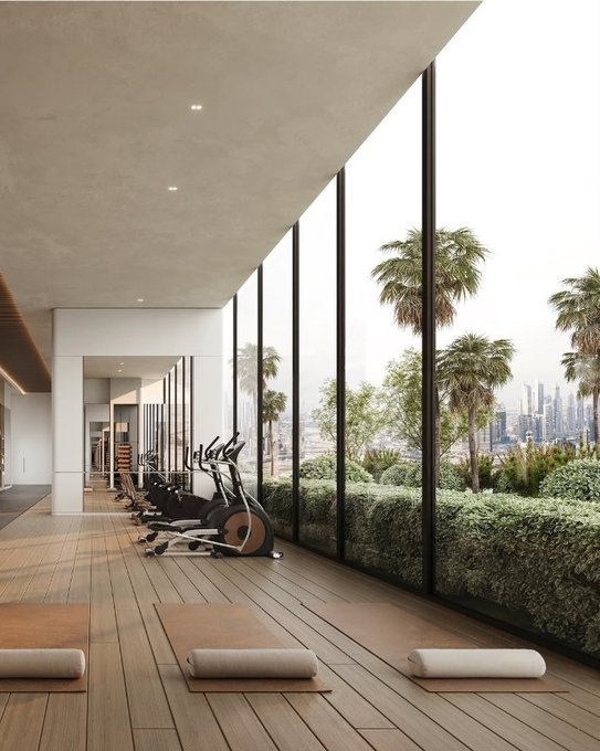 A modern gym with floor-to-ceiling windows overlooking palm trees and a city skyline, featuring stationary bikes and yoga mats on wooden flooring.