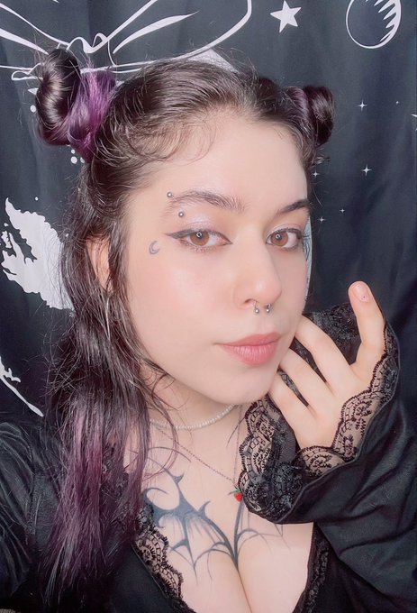 A portrait of a young woman with dark and purple hair styled in two buns, featuring facial piercings, eye makeup, and gothic-style clothing, posing in front of a dark tapestry with celestial designs.
