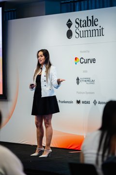 The image shows a woman standing on stage, giving a presentation or speech. She appears to be wearing a black dress and white jacket. The background features a large screen displaying the words "Stable Summit" and "MIDAS". There are also several people in the audience, some of whom are sitting while others stand. A laptop can be seen placed on a table near the stage.