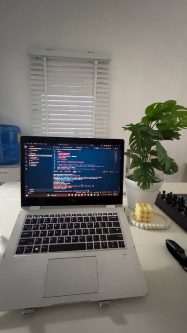 A laptop on a white desk displays lines of code in a dark-themed editor, accompanied by a potted Monstera plant and a keyboard.