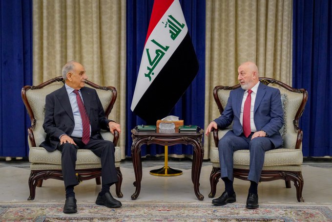 Two men in suits sit in armchairs in a formal meeting setting, with the Iraqi flag (الله أكبر) displayed between them and blue and gold curtains in the background.
