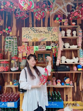 A woman in a white dress stands inside a colorful Vietnamese souvenir shop, holding a decorative item, with shelves of merchandise including '2-7UP' Pepsi products and signs reading 'Heaven Peace' visible behind her.