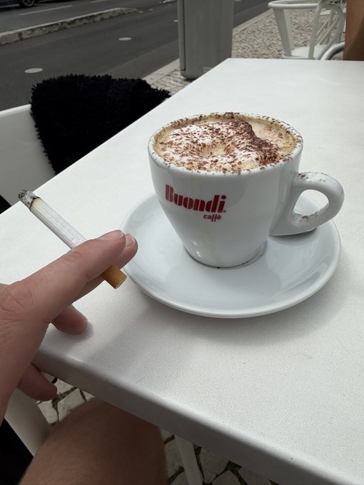 A hand holding a cigarette rests on a white café table next to a cappuccino in a 'Buondi caffe' branded cup, topped with cinnamon and cocoa powder.