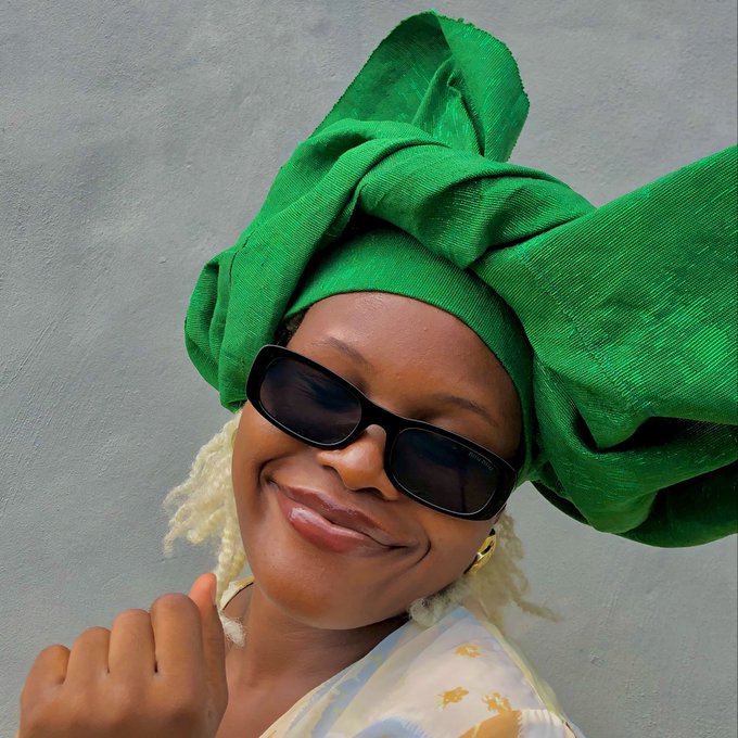 A smiling woman wearing a large green headwrap, sunglasses, and a patterned top gives a thumbs-up against a plain wall.