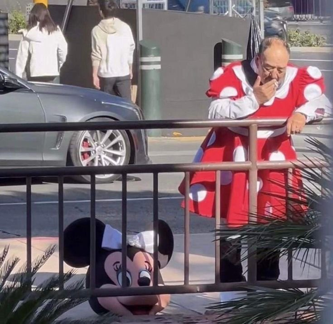 A man dressed in a red and white polka-dot clown costume leaning on a metal railing and smoking, with a Mickey Mouse mascot sitting on the ground nearby.