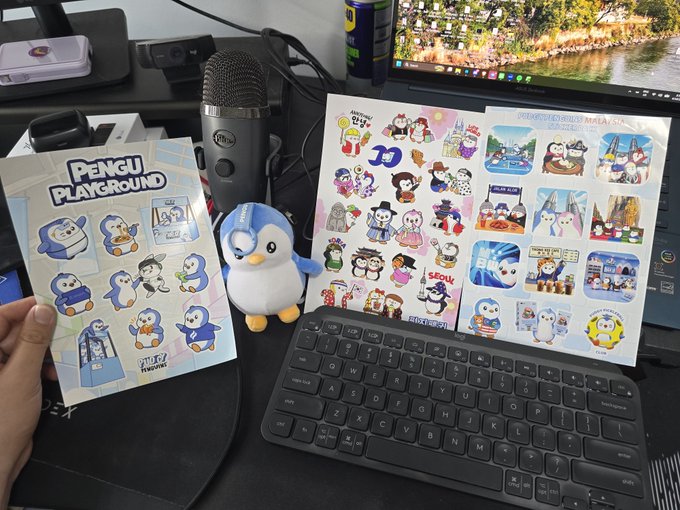 A desk setup featuring a keyboard, microphone, and a monitor, with a person displaying a 'Pengu Playground' booklet and a sheet of character stickers alongside a small plush toy.