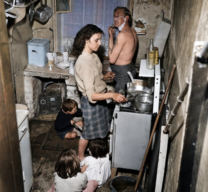 A vintage photograph depicts a family in a small, rustic kitchen, where a man is shaving, a young woman cooks at the stove, and children sit on the floor.