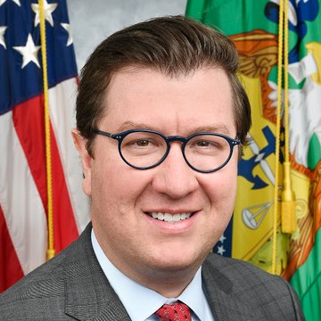 A headshot of a smiling man wearing glasses and a suit, with an American flag and another flag visible in the background.