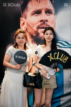 Two women pose at an XClub event in front of a large Messi promotional poster, holding XClub-branded signs and a folding fan.