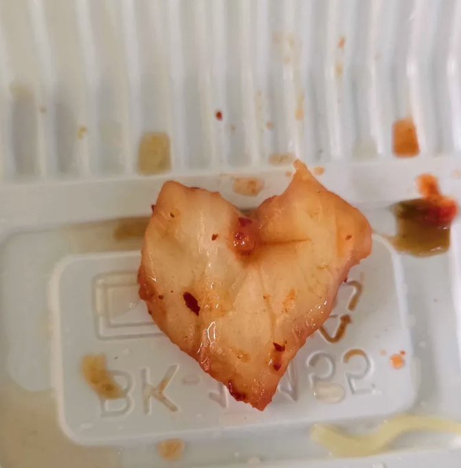 The image features a heart-shaped piece of food that appears to be a type of meat or fish, possibly chicken wings. The food has sauce dripped from it, indicating that it was cooked recently. The background of the image shows a white plastic container with sauce on it and some sauce residue around the edges. There is also text in the image that reads "A day full of love with Tria! @useTria's Dokdo DAO campaign is wrapping up on June 1st!".
