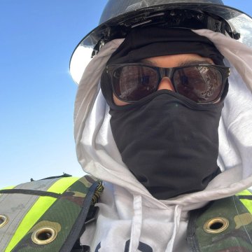 A close-up selfie of a person wearing a hood, black face mask, and dark sunglasses, with a reflective metallic visor or helmet visible above.
