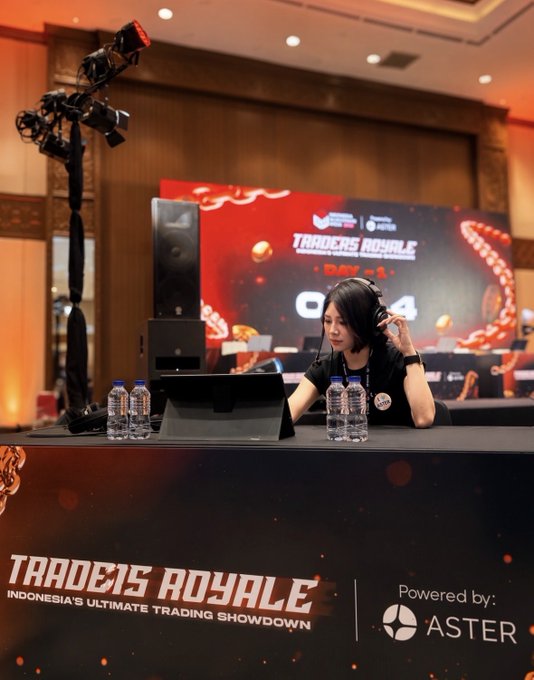 A woman wearing headphones sits at a competition desk for 'Traders Royale - Indonesia's Ultimate Trading Showdown' powered by Aster, with a camera and large display behind her.