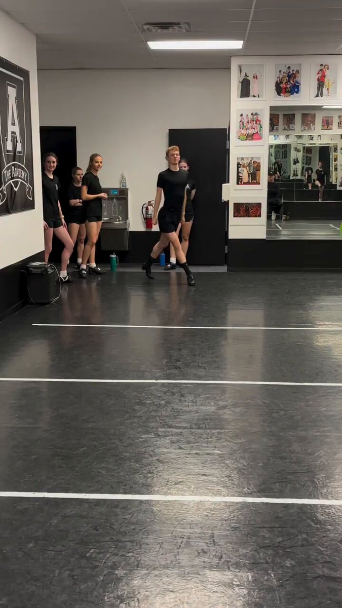The image shows a group of people standing in a dance studio, with some of them wearing black outfits. They are all facing away from the camera, giving an impression that they might be posing for a photo or waiting for their turn to perform on stage. The room is filled with mirrors and has a large poster hanging on one of the walls. There's also a suitcase placed in the middle of the floor, which could possibly belong to one of the dancers.