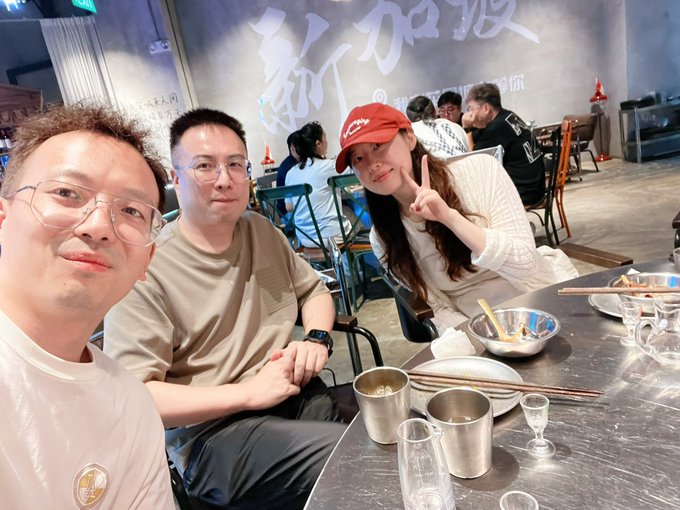 Group selfie taken at a restaurant showing three people – two men in the foreground and a woman in a red cap making a peace sign – seated at a table with bowls, cups, and chopsticks, with Chinese calligraphy on the wall behind them.
