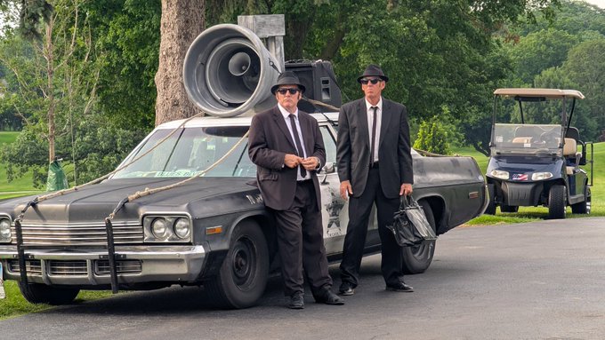 Two men in black suits and hats standing in front of a black vintage car with large speakers mounted on top, parked on a road with a golf cart and greenery in the background.
