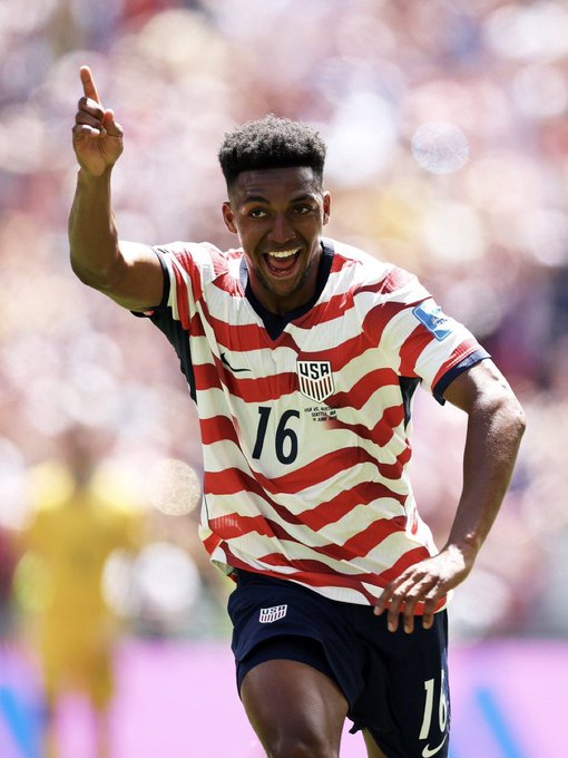 The image features a soccer player wearing an American flag-themed jersey, with the number 16 prominently displayed. He appears to be celebrating his success or victory during a match by giving a high five gesture. The background of the photo shows other players on the field, indicating that this is a competitive game being played.