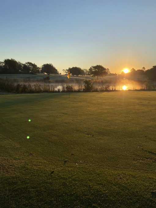 A scenic sunset view over a golf course shows a yellow flag on the green in the foreground, with a pond reflecting the setting sun and trees lining the horizon.