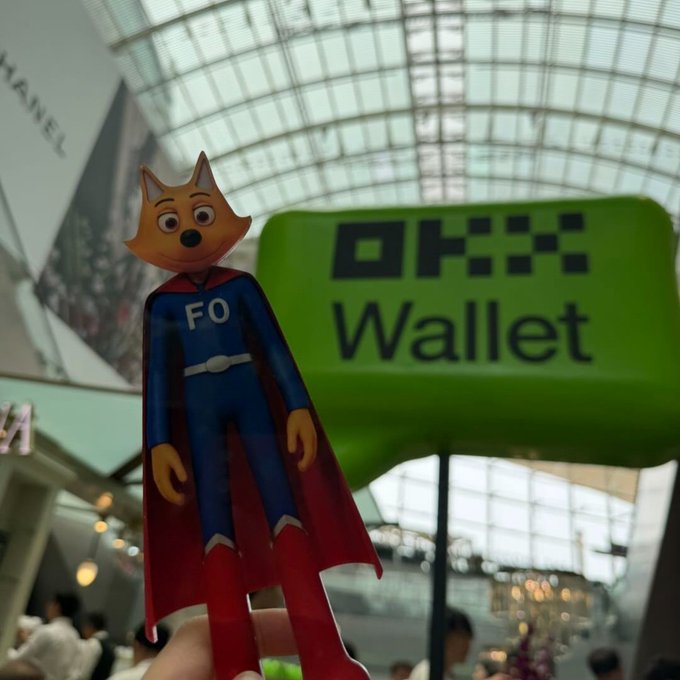 A close-up of a hand holding a small Zorro-style fox action figure wearing a blue outfit with "FO" on the chest, standing next to a green "OKX Wallet" sign in a mall.