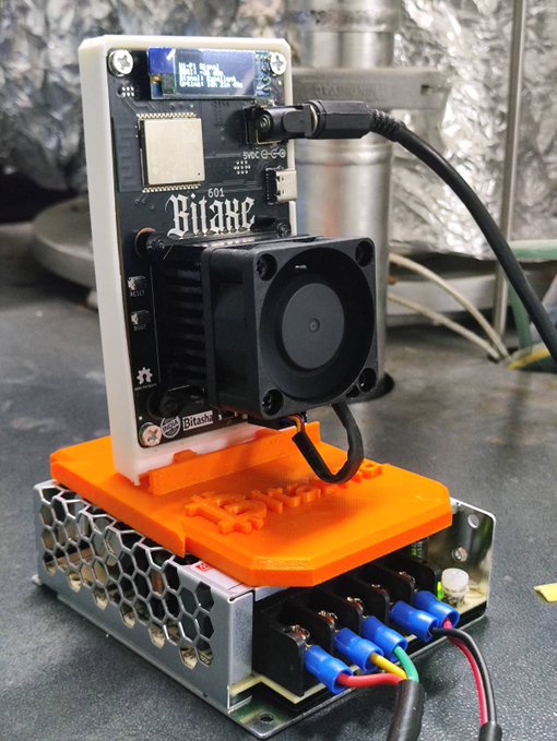 A Bitaxe 601 Bitcoin solo miner device with a cooling fan and small display screen, mounted on an orange 3D-printed base attached to a power supply unit.