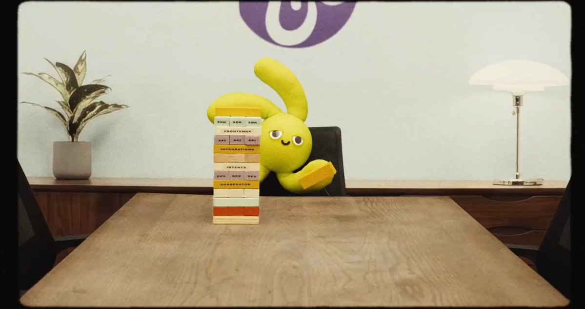 A yellow plush toy character sits on a wooden table next to a tall stack of Jenga-like blocks labeled with words like 'SDK', 'FRONTEND', 'API', 'INTEGRATIONS', 'INTENTS', 'DEX', and 'AGGREGATOR', with a plant and lamp visible in the background.