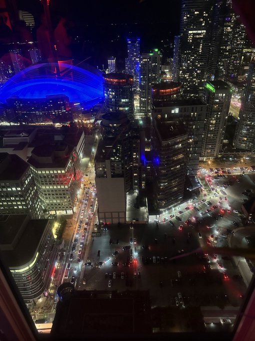 A nighttime cityscape view from a high window, showing illuminated skyscrapers, a stadium with blue lighting, and traffic on the streets.