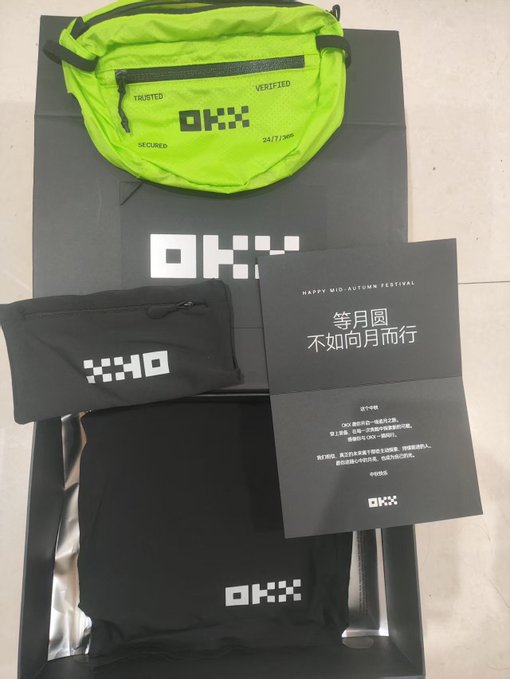 A promotional box containing a neon green fanny pack, black pouches, and a card with the 'OKX' logo. Text on the card includes 'HAPPY MID-AUTUMN FESTIVAL', '等月圆 不如向月而行' (Waiting for the full moon, better to walk toward the moon), and 'TRUSTED VERIFIED SECURED 24/7/365'.