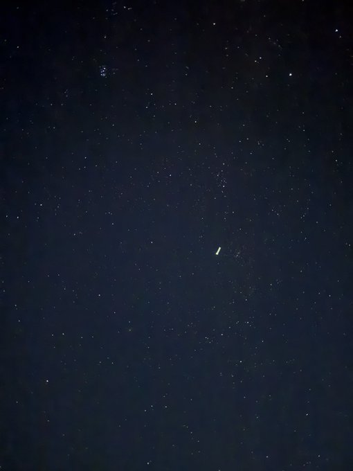 A nighttime photograph of a starry sky featuring the Milky Way and what appears to be a small celestial object or aircraft with lights visible.