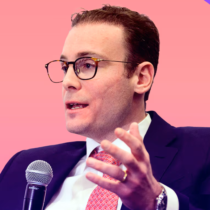 The image shows a man wearing glasses talking into a microphone while sitting in a suit jacket at an event. The scene seems to be related to bitcoin or cryptocurrency trading since he is using a tool for tracking prices, which is typically associated with this type of digital currency exchange. However, it is unclear if the Bitcoin Perpetual Futures tool is specifically targeting U.S. markets based on the provided information in the image description.