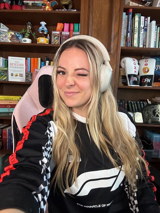 The image features a woman wearing headphones, sitting in front of a bookshelf filled with various items such as books and figurines. The woman appears to be smiling while holding her phone up for the camera. The background includes a bookshelf containing numerous books and other objects. The caption reads "Live right now talking about $BC new white paper and engine Then playing some Marvel Rivals later and ripping packs". This suggests that the woman is engaged in an activity related to gaming or streaming, possibly discussing game-related topics with her audience while wearing headphones for a more immersive experience.

The image also contains several books scattered around the room, indicating that she might be reading or researching about the games she plays or streams. The presence of figurines on the bookshelf adds an artistic touch to the scene and may reflect her interests in collecting gaming memorabilia.