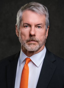 A distinguished-looking middle-aged man with silver-gray hair and a matching full beard is shown in a professional headshot against a dark, textured background. He wears a dark navy suit jacket, a crisp white dress shirt, and a vibrant orange tie that pops against his attire. His expression is serious and contemplative, with piercing eyes gazing directly at the camera, suggesting an air of authority and confidence. The image likely represents Michael Saylor, the founder and executive chairman of MicroStrategy (now known as Strategy), as referenced in the accompanying post about the company's latest Bitcoin acquisition of 950 BTC for approximately $80 million.