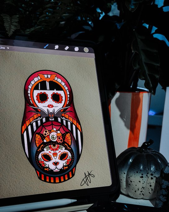 A digital tablet displays a vibrant Day of the Dead-themed illustration of a sugar skull matryoshka doll, sitting next to a small black pumpkin and surrounded by dark foliage.