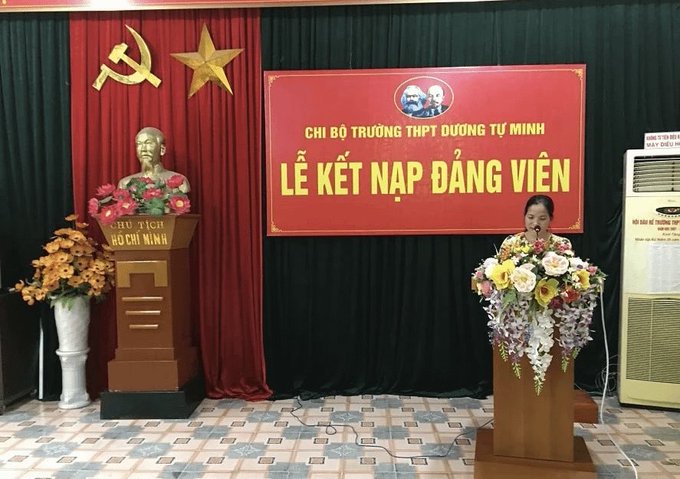 A woman speaks at a podium during a Vietnamese ceremony, with a red banner reading 'LỄ KẾT NẠP ĐẢNG VIÊN' and a bust of Ho Chi Minh nearby.