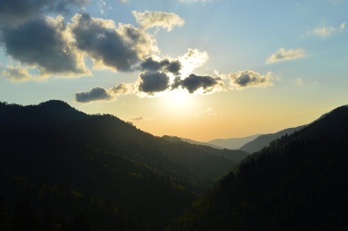 A scenic landscape photograph of the sun setting behind forested mountains under a sky scattered with clouds.
